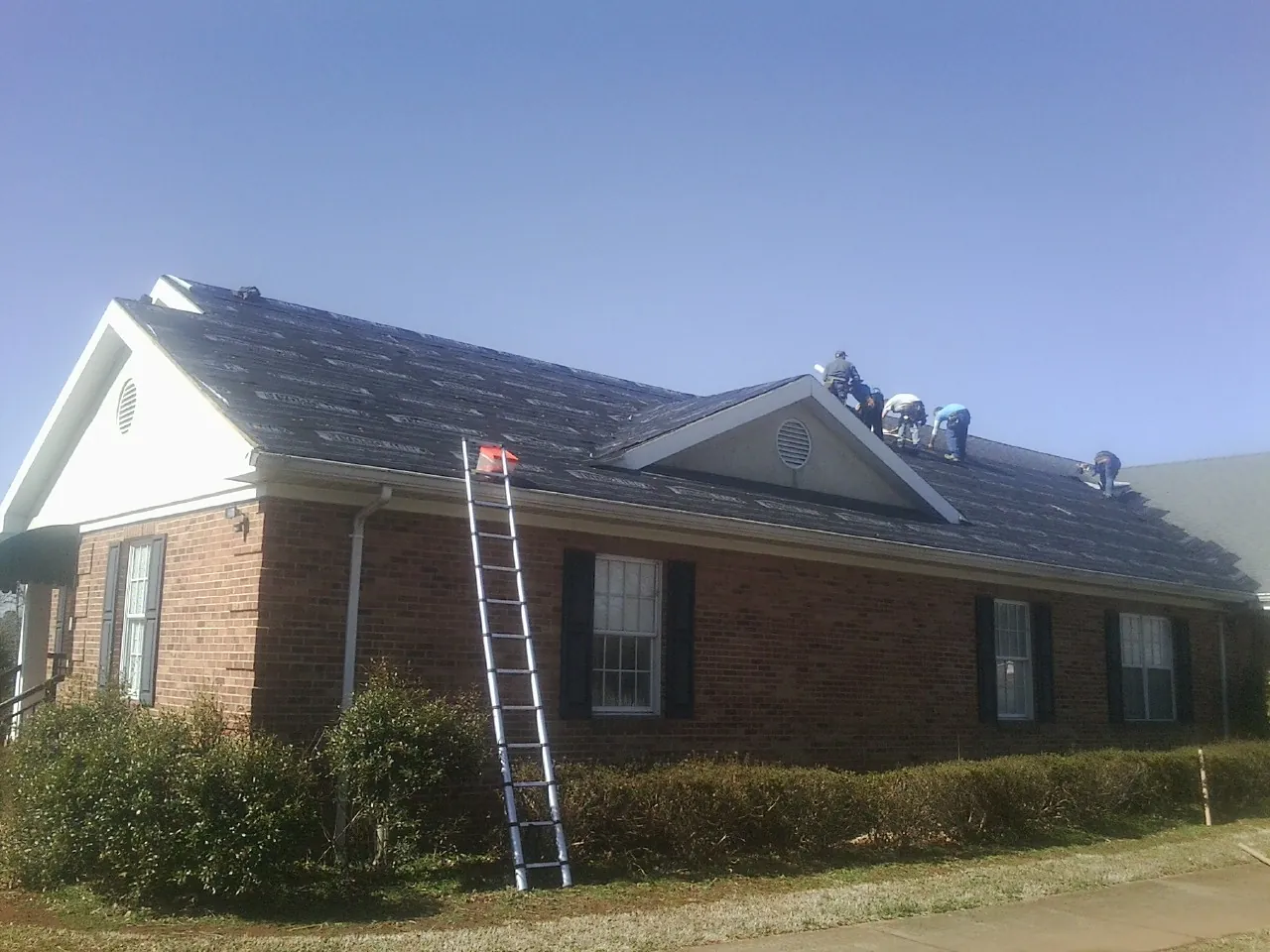 Crew tearing shingles off a brick ranch roof with a ladder set — Metal Roof Repair work by our crew in Ramer