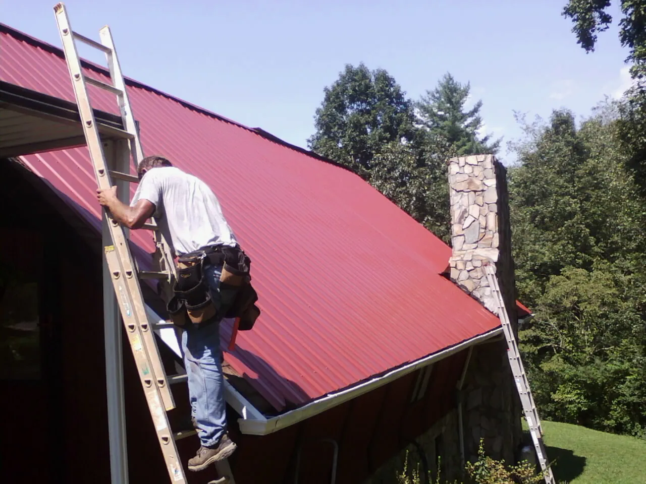 Roofer on a ladder beside a red metal roof and stone chimney — Soffit & Fascia Repair by Roofing Brothers in Ramer